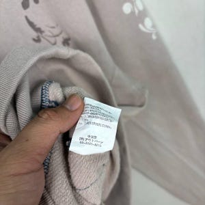 May include: A close-up of a light mauve sweatshirt with a floral pattern. The garment has a label with Japanese text and the phone number 03-3891-8215. The sweatshirt's fabric is visible, and the stitching is detailed.