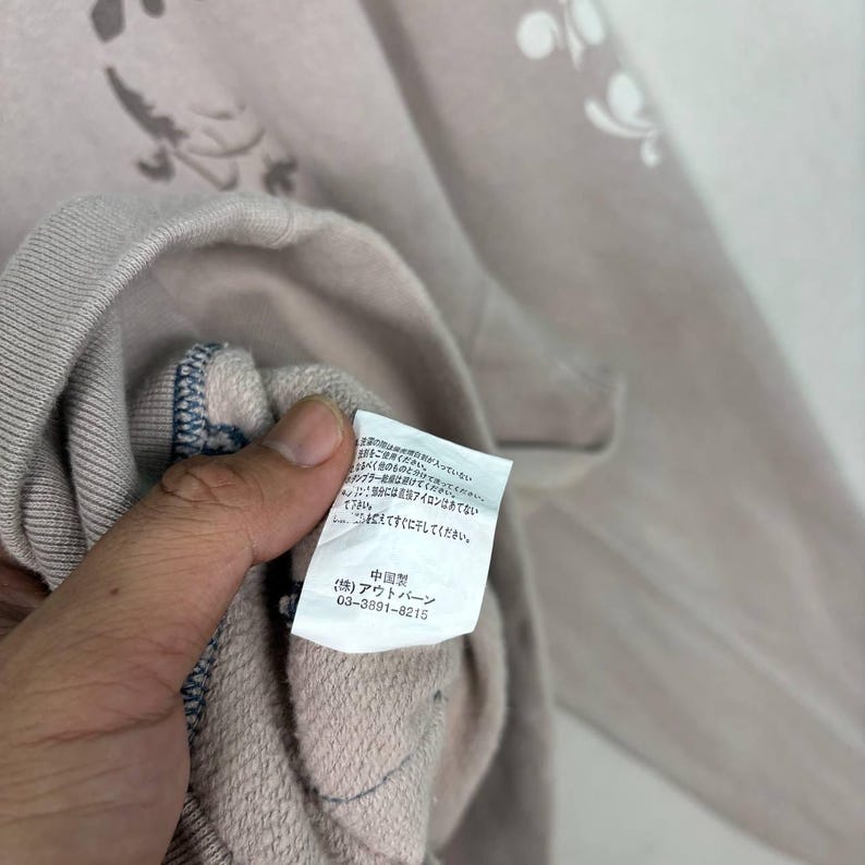 May include: A close-up of a light mauve sweatshirt with a floral pattern. The garment has a label with Japanese text and the phone number 03-3891-8215. The sweatshirt's fabric is visible, and the stitching is detailed.