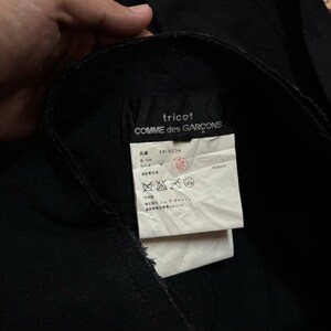 May include: Close-up of a black garment with a label. The label reads "tricot COMME des GARCONS" and includes Japanese text and symbols. The garment appears to be made of a dark fabric, possibly wool or a similar material.