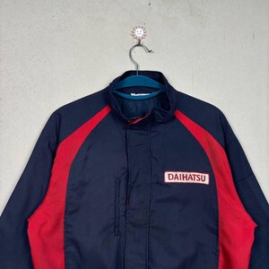 May include: Navy blue jacket with red accents and the "DAIHATSU" logo. The jacket has a stand-up collar and a full-length zipper. The jacket is hanging on a metal hanger.