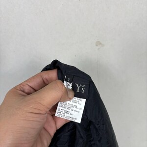 Op de afbeelding: Een zwart kledinglabel met de merknaam "Y's" in het wit. Het label bevat onderhoudsinstructies en een maatindicator. De stof lijkt een donker, mogelijk nylon materiaal te zijn.