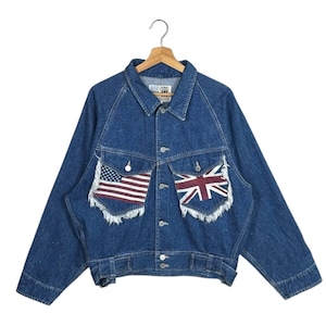 May include: A blue denim jacket with long sleeves and a button-up front. The jacket features two front pockets, each adorned with a flag design: the American flag on the left and the Union Jack on the right. The edges of the pockets have a frayed design.