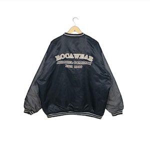 Może przedstawiać: Czarna i szara kurtka bomberka z napisem "BOCAWEAR APPAREL COMPANY EST. 1999" wyhaftowanym na plecach. Kurtka ma prążkowany kołnierz, mankiety i dół z białymi paskami.