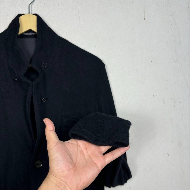 Może przedstawiać: Czarny wełniany płaszcz z kołnierzem mandaryńskim i zapięciem na guziki. Płaszcz ma jednorzędowy kr&oacute;j i składany mankiet. Wisi na drewnianym wieszaku.