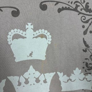 May include: Close-up of a light gray fabric with a repeating crown design in white. The crowns have a kangaroo logo. Dark gray floral patterns are in the upper right corner. There is a brown stain on the fabric.