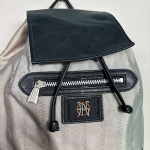 Op de afbeelding: Een zilveren rugzak met een zwarte leren flap en accenten. De rugzak heeft een zwart ritsvak, een zwart leren patch met een logo en zwarte trekkoordsluitingen. De ritssluiting heeft een decoratief ontwerp.