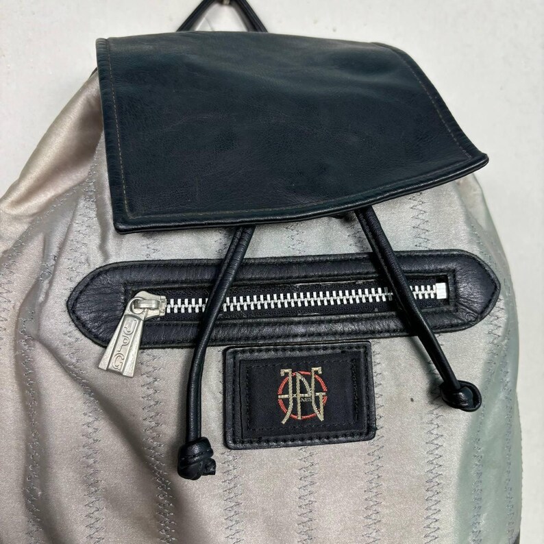 Op de afbeelding: Een zilveren rugzak met een zwarte leren flap en accenten. De rugzak heeft een zwart ritsvak, een zwart leren patch met een logo en zwarte trekkoordsluitingen. De ritssluiting heeft een decoratief ontwerp.