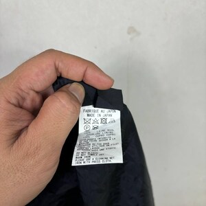 Op de afbeelding: Een zwart kledinglabel met witte tekst en verzorgingssymbolen. Het label luidt "FABRIQUE AU JAPON MADE IN JAPAN" en bevat de stofsamenstelling en wasvoorschriften. Het label wordt door een hand tegen een lichte achtergrond gehouden.