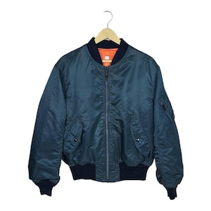 May include: A teal bomber jacket with a navy blue collar, cuffs, and waistband. The jacket has a zipper closure, two front pockets, and a small pocket on the left sleeve. The interior lining is orange. The jacket is hanging on a wooden hanger.