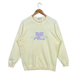 Pode incluir: Uma camisola amarelo p&aacute;lido com a palavra "courreges" bordada num design lil&aacute;s claro e azul. A camisola est&aacute; pendurada num cabide de madeira.