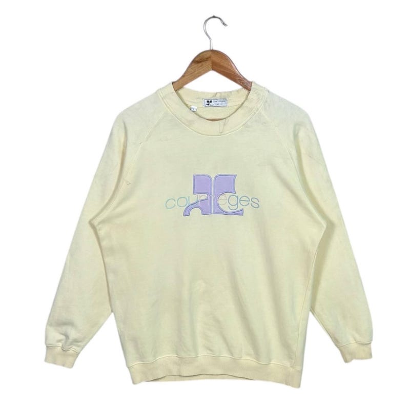Pode incluir: Uma camisola amarelo p&aacute;lido com a palavra "courreges" bordada num design lil&aacute;s claro e azul. A camisola est&aacute; pendurada num cabide de madeira.