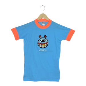 May include: A light blue t-shirt with orange trim on the collar and sleeves. The shirt features a cartoon graphic of a smiling face with the text "squeeze me, I'll talk to you!" The shirt is hanging on a wooden hanger.