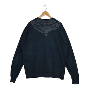 Könnte beinhalten: Schwarzes Sweatshirt mit einem grafischen Design auf dem oberen Rücken. Das Design zeigt ein stilisiertes Bild einer Schutzbrille und einer Gesichtsbedeckung. Das Sweatshirt hängt an einem Holzbügel vor weißem Hintergrund.