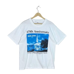 May include: A white short-sleeved t-shirt featuring a blue graphic print of a church building. The text reads 175th Anniversary 1824-1999 Trinity United Methodist Church Tallahassee, Florida. The shirt is displayed on a wooden hanger against a white background.