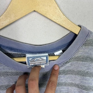 May include: A light blue and gray striped crew neck shirt with a small logo tag. The shirt is hanging on a wooden hanger. The tag has the brand name and logo.
