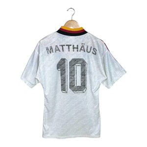 May include: White football shirt with the name "MATTH&Auml;US" and the number "10" printed on the back. The shirt has a patterned design and a collar with black, red, and gold accents. The shirt is hanging on a wooden hanger.