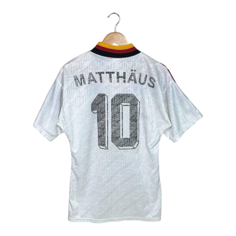 May include: White football shirt with the name "MATTH&Auml;US" and the number "10" printed on the back. The shirt has a patterned design and a collar with black, red, and gold accents. The shirt is hanging on a wooden hanger.