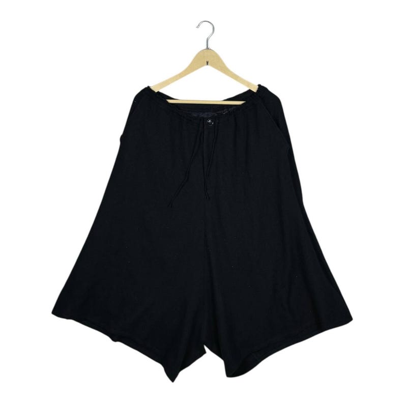 Op de afbeelding: Zwarte shorts met wijde pijpen, hangend aan een houten hanger. De shorts hebben een taille met trekkoord en een knoopsluiting. De stof lijkt een zacht, vloeiend materiaal te zijn.