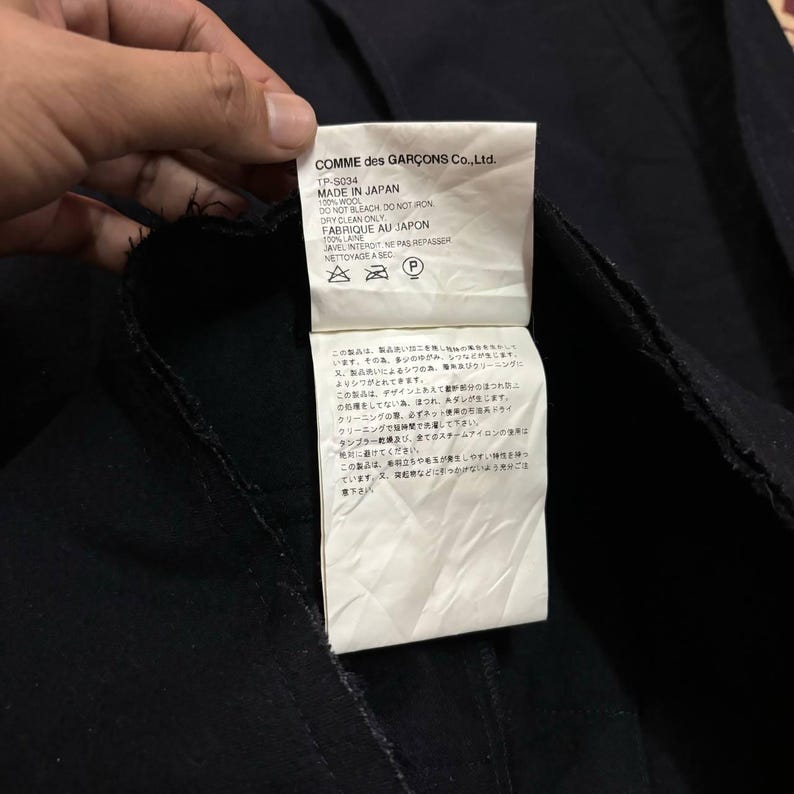 May include: A black garment with a white tag attached. The tag features the text "COMME des GAR&Ccedil;ONS Co.,Ltd." and "MADE IN JAPAN." The tag also includes care instructions and fabric composition details, including "100% WOOL."
