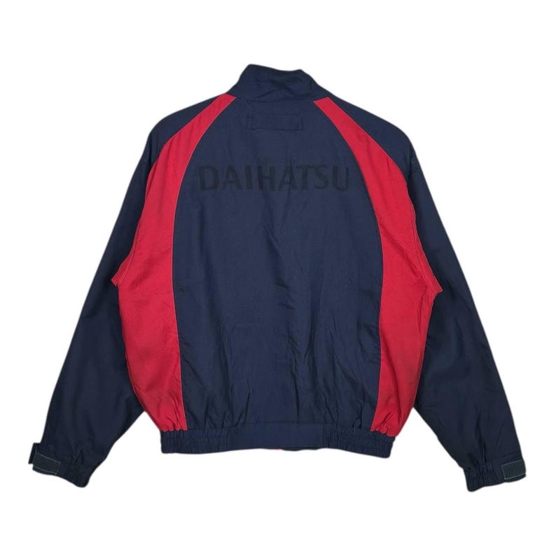May include: Navy blue and red jacket with the word "DAIHATSU" printed across the back. The jacket has a stand-up collar, long sleeves, and a zipper closure. The sleeves have adjustable cuffs.