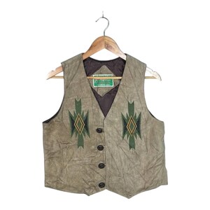 Peut inclure: Un gilet en daim beige avec une doublure intérieure marron foncé et quatre boutons sur le devant. Le devant présente deux motifs géométriques en losange vert et noir symétriques. Une étiquette rectangulaire verte avec du texte blanc est cousue.