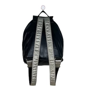 Op de afbeelding: Zwarte rugzak met zilverkleurige bandjes en accenten. De rugzak heeft een afgeronde vorm en een handvat aan de bovenkant. De bandjes zijn verstelbaar en lijken van satijnachtig materiaal te zijn. De rugzak hangt tegen een witte achtergrond.