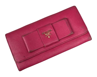 May include: A magenta Prada wallet with a bow detail. The wallet features a rectangular shape with a flap closure and a bow-shaped accent in the center. The Prada logo is visible on the bow. The wallet is made of a textured material.