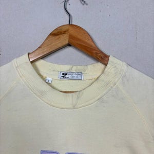 Pode incluir: Uma camisola amarela p&aacute;lida com gola redonda e mangas compridas, pendurada num cabide de madeira. A etiqueta diz "courr&egrave;ges and co." e a palavra "DC" &eacute; bordada em lavanda na frente.