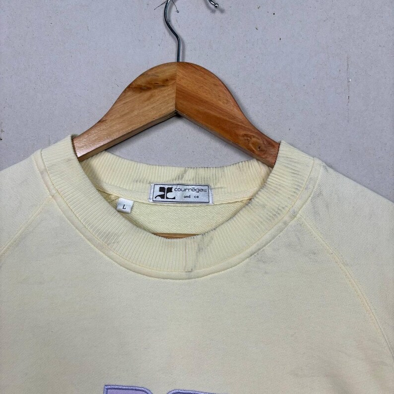 Pode incluir: Uma camisola amarela p&aacute;lida com gola redonda e mangas compridas, pendurada num cabide de madeira. A etiqueta diz "courr&egrave;ges and co." e a palavra "DC" &eacute; bordada em lavanda na frente.