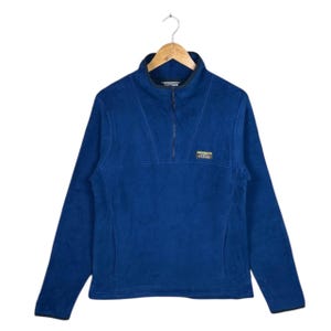May include: A blue fleece pullover with a half-zip closure and a small logo patch. The long-sleeved top has a high collar and is hanging on a wooden hanger. The fleece material appears soft and warm, suitable for cooler weather.
