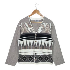 May include: A button-up cardigan with a V-neck and long sleeves. The sweater features a mix of patterns, including geometric and paisley designs in shades of gray, black, white, and brown. The sleeves are a solid gray color.