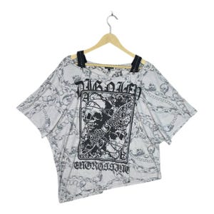 Op de afbeelding: Een lichtgrijs t-shirt met een schedel- en kettingpatroon. Het shirt heeft een grote afbeelding van schedels en de woorden "Pirated" en "Engrossing" in het zwart. Het shirt heeft zwarte bandjes en korte mouwen.