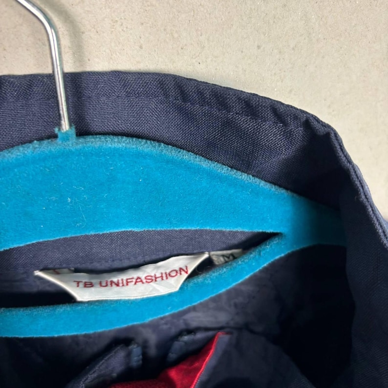 May include: Close-up of a navy blue garment hanging on a teal blue hanger. The garment has a label that reads "TB UNIFASHION". The garment's interior lining is red. The hanger is suspended against a light gray background.