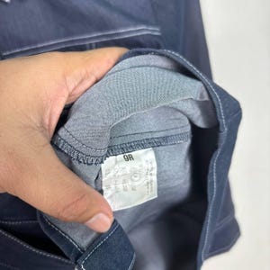 May include: Close-up of a dark blue denim garment, possibly a shirt or jacket, with visible stitching and a fabric tag. The tag displays text and numbers, including "9R". The garment is held by a hand.