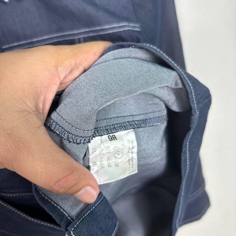 May include: Close-up of a dark blue denim garment, possibly a shirt or jacket, with visible stitching and a fabric tag. The tag displays text and numbers, including "9R". The garment is held by a hand.