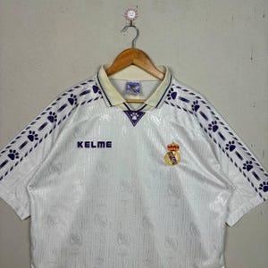 K&ouml;nnte beinhalten: Wei&szlig;es Fu&szlig;balltrikot mit lila Pfotenabdruck-Akzenten auf den &Auml;rmeln und am Kragen. Mit dem "KELME"-Logo und einem Vereinswappen. Das Trikot h&auml;ngt an einem Holzkleiderb&uuml;gel.