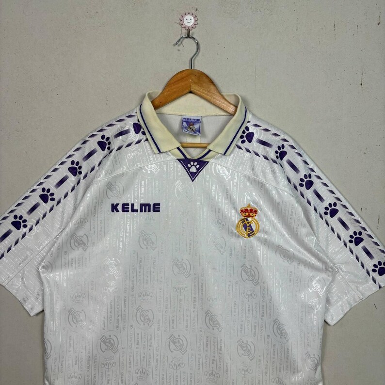 K&ouml;nnte beinhalten: Wei&szlig;es Fu&szlig;balltrikot mit lila Pfotenabdruck-Akzenten auf den &Auml;rmeln und am Kragen. Mit dem "KELME"-Logo und einem Vereinswappen. Das Trikot h&auml;ngt an einem Holzkleiderb&uuml;gel.
