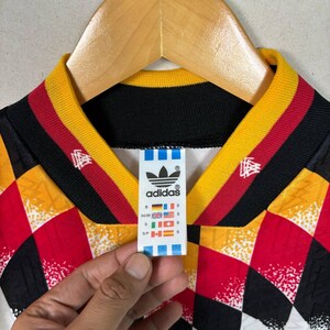 May include: Vintage Adidas football shirt with a geometric pattern in red, yellow, black, and white. The collar has black, yellow, and red stripes. The Adidas logo and size tag are visible.
