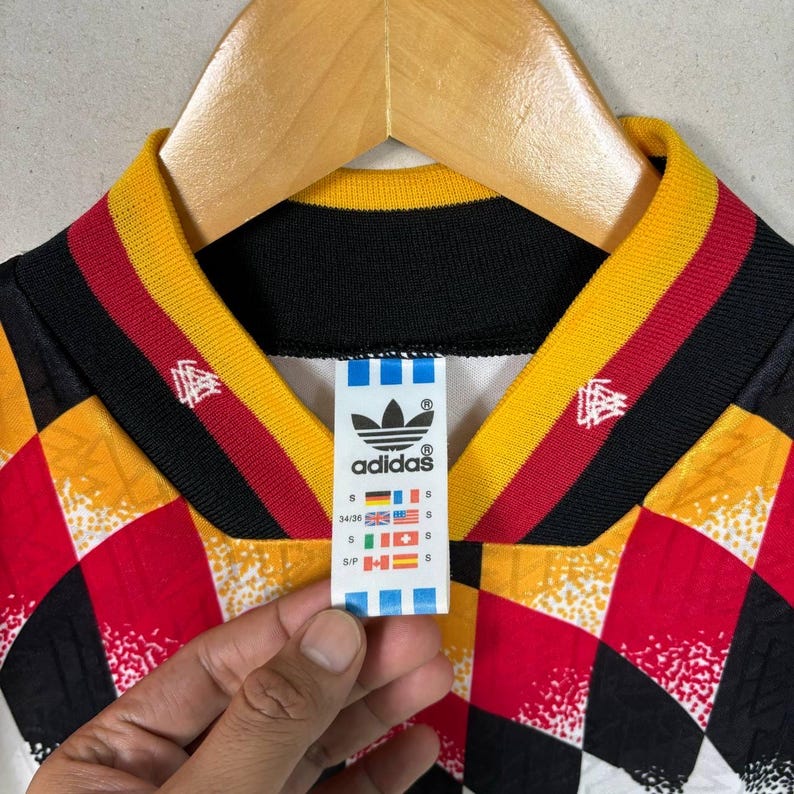 May include: Vintage Adidas football shirt with a geometric pattern in red, yellow, black, and white. The collar has black, yellow, and red stripes. The Adidas logo and size tag are visible.