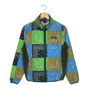 May include: A zip-up jacket with a patchwork design featuring paisley patterns in shades of blue, green, and brown. The jacket has a green collar, zipper, and side panels. A small label with text is on the chest.