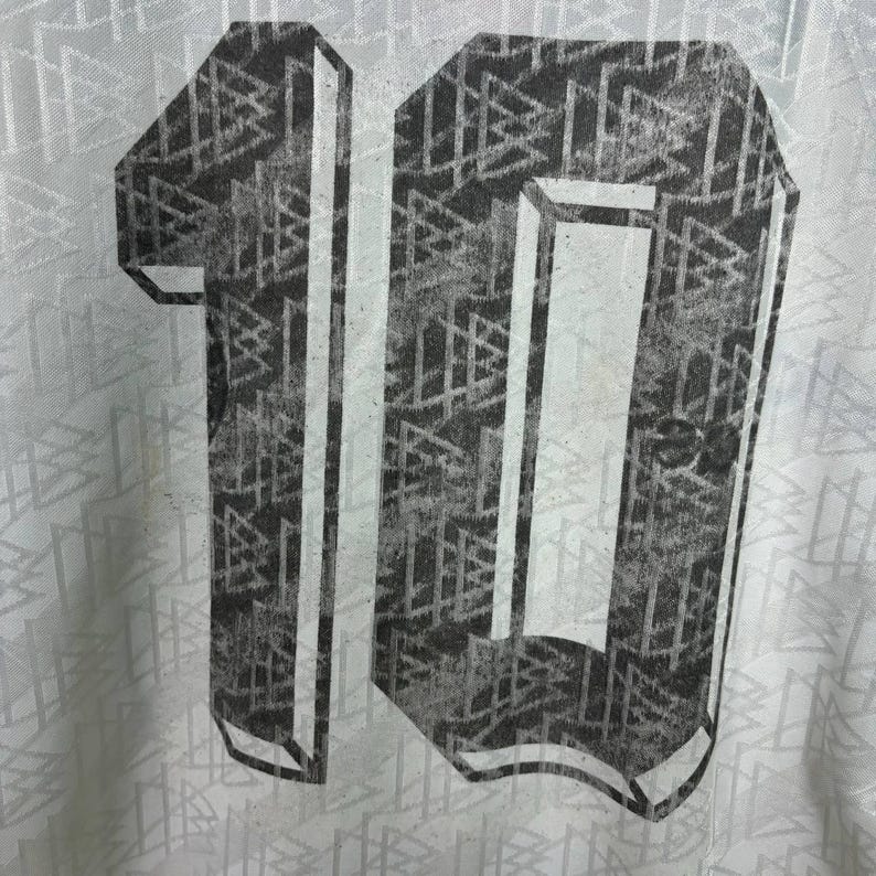May include: Close-up of a white jersey featuring a large, distressed number 10 on the back. The number is grey with a geometric pattern. The jersey has a subtle, repeating pattern.