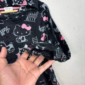 Puede incluir: Sudadera con capucha negra con un patr&oacute;n repetido de personajes de Hello Kitty con lazos rosas y el texto "Hello Kitty". La sudadera est&aacute; hecha de un material suave y afelpado.