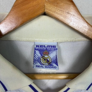 K&ouml;nnte beinhalten: Cremefarbenes Fu&szlig;balltrikot mit blauen Akzenten, an einem Holzkleiderb&uuml;gel aufgeh&auml;ngt. Das Etikett des Trikots zeigt "KELME SPONSOR TECNICO" und das Real Madrid Logo.