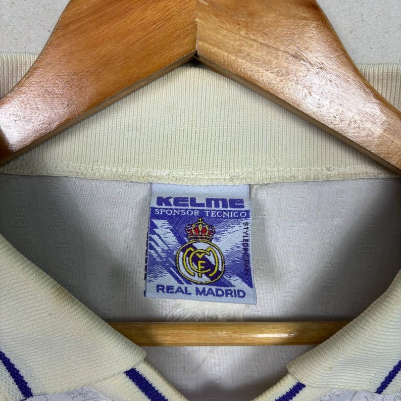 K&ouml;nnte beinhalten: Cremefarbenes Fu&szlig;balltrikot mit blauen Akzenten, an einem Holzkleiderb&uuml;gel aufgeh&auml;ngt. Das Etikett des Trikots zeigt "KELME SPONSOR TECNICO" und das Real Madrid Logo.