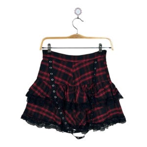 May include: Red and black plaid skirt with a layered ruffle design and black lace trim. The skirt has grommet details and is hanging on a wooden hanger.