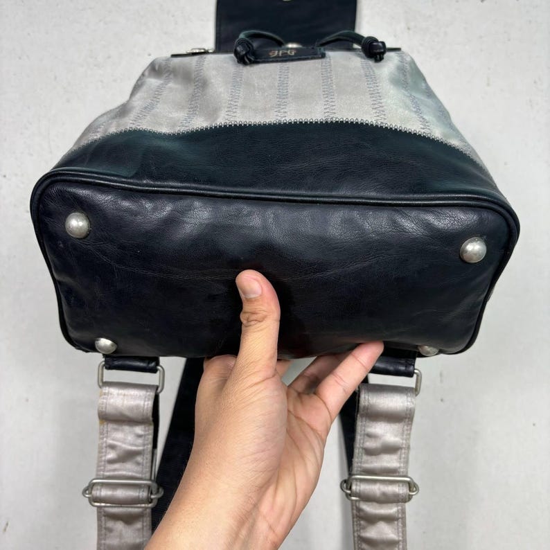 Op de afbeelding: Een zwarte en zilveren rugzak met een leren basis en een gestreepte stoffen bovenkant. De rugzak heeft zilverkleurige hardware, verstelbare bandjes en een kleine bovenklep. Het woord "arg" is bovenaan zichtbaar.