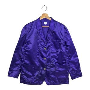 Puede incluir: Una vibrante blazer morada con acabado satinado, con cuello con solapas clásico, tres botones visibles y dos bolsillos delanteros. La chaqueta está colgada de una percha de madera, mostrando su diseño a medida y su textura suave.