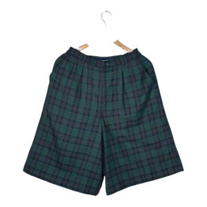 May include: Green and navy plaid shorts hanging on a wooden hanger. The shorts have a button closure and pleats at the waist. The fabric has a classic plaid pattern in shades of green and navy.