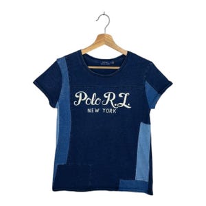 Puede incluir: Una camiseta de mezclilla azul oscuro con mangas cortas, con un diseño de patchwork. La camiseta muestra el texto "Polo R.L. NEW YORK" en escritura blanca en la parte delantera. La camiseta está colgada en una percha de madera.