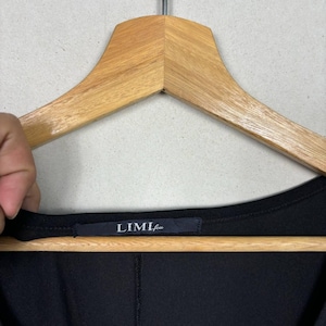 May include: Black garment with a visible label that reads "LIMI feu". The label is rectangular and dark, with white text. The garment is hanging on a wooden hanger.